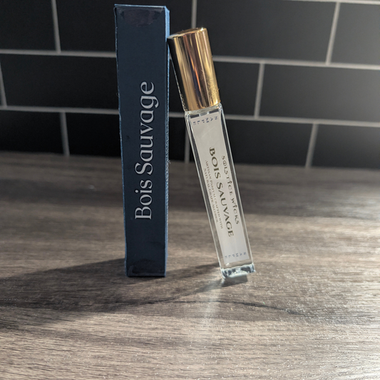 A clear glass perfume sample vial with a metallic gold cap stands next to a tall dark blue box labeled Bois Sauvage on a wooden tabletop, with a dark tiled wall in the background.