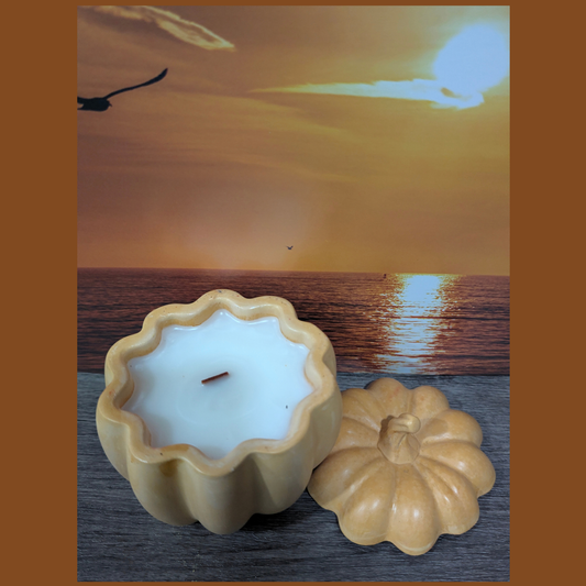A candle set with a scalloped cream-colored handcrafted pumpkin and a matching pumpkin-shaped lid, displayed on a dark surface against a sunset ocean backdrop.
