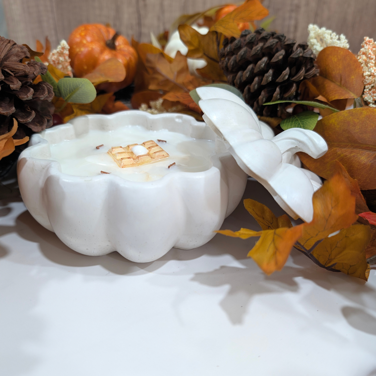 the front of a handcrafted White large pumpkin front of a falls decoration like leaves, pine corn. white flower inside of the pumpkin is fill with coconut apricot wax and have a waffle decoration