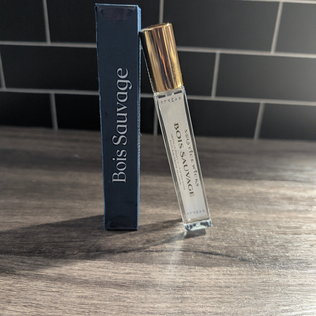 A clear glass perfume sample vial with a metallic gold cap stands next to a tall dark blue box labeled Bois Sauvage on a wooden tabletop, with a dark tiled wall in the background.
