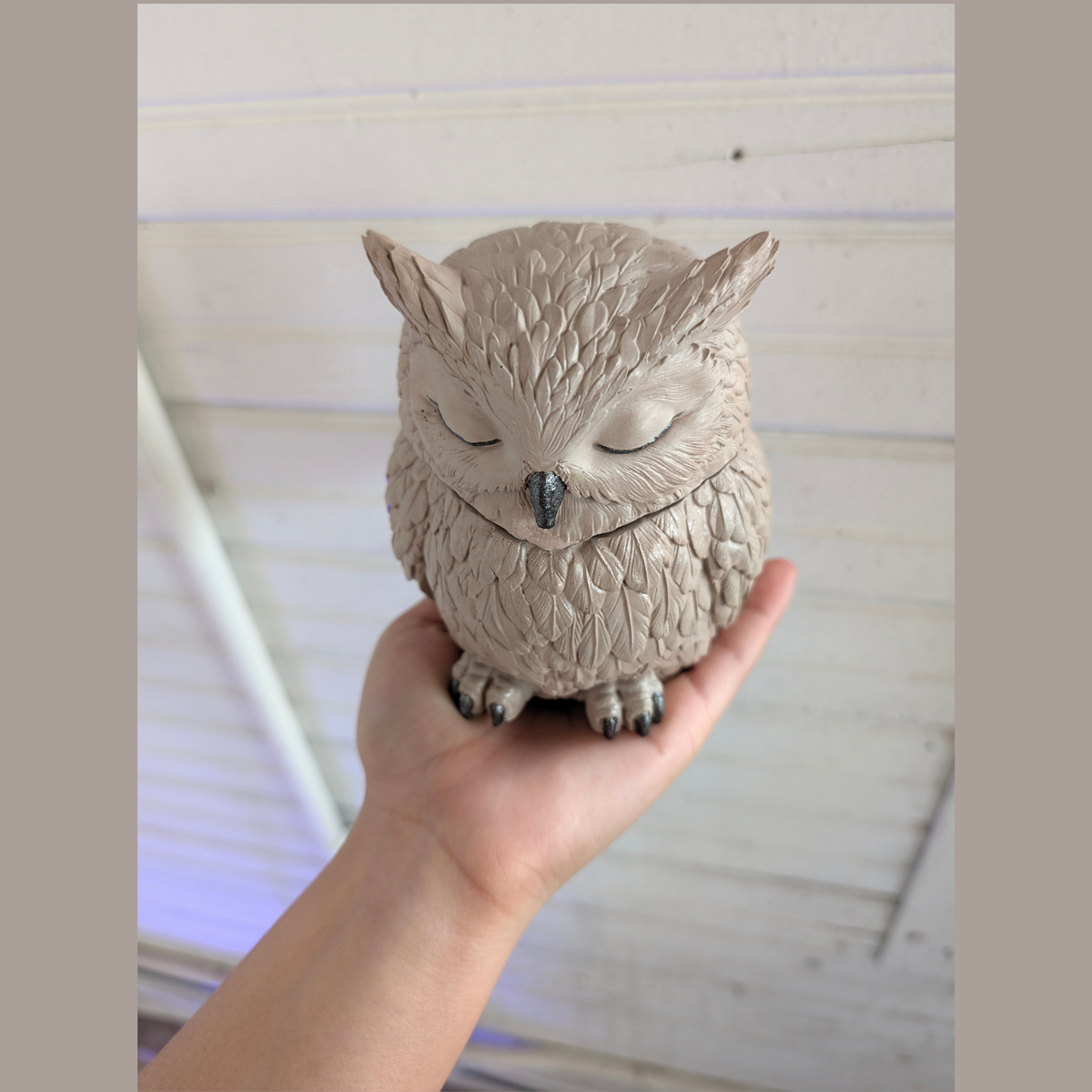 Handcrafted Owl the color of the Owl are super light brown the owl is in top of a hand  in front of a white wood wall