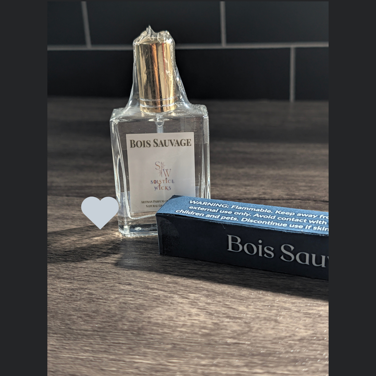 A bottle of Bois Sauvage cologne by Solstice Wicks, wrapped in protective plastic, standing next to its dark blue cardboard packaging box on a dark wood surface.