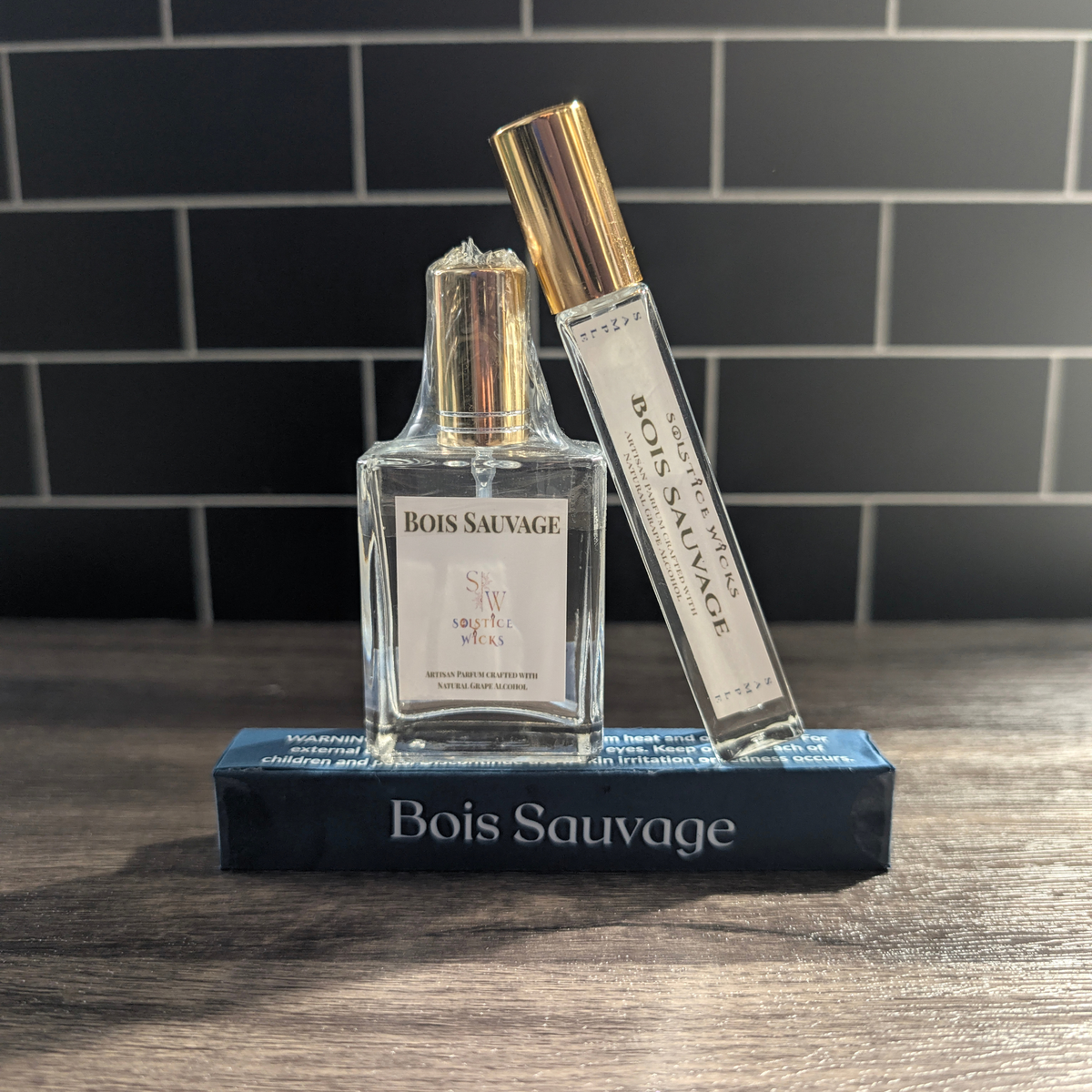 A bottle of Bois Sauvage cologne and a matching To-Go spray stand on top of a dark blue box on a wooden surface, with a dark tiled background.