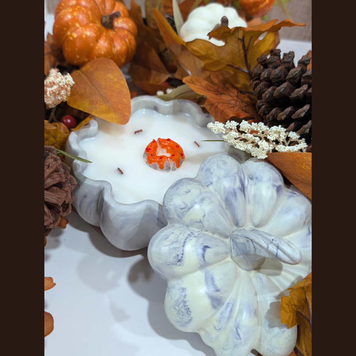 A decorative white candle in a handcrafted pumpkin-shaped vessel, surrounded by autumn leaves, pinecones, and a small orange pumpkin on a tabletop.