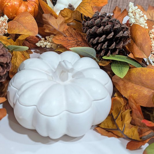 We have a handcrafted White large pumpkin in front of a falls decoration like leaves, pine corn. white flower in top of a white countertop