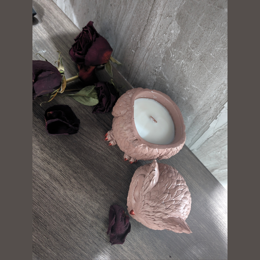 Handcrafted owl-shaped concrete candle  in a dusty brown color,  open to reveal a white candle inside  on a gray wooden floor with dried dark purple roses.