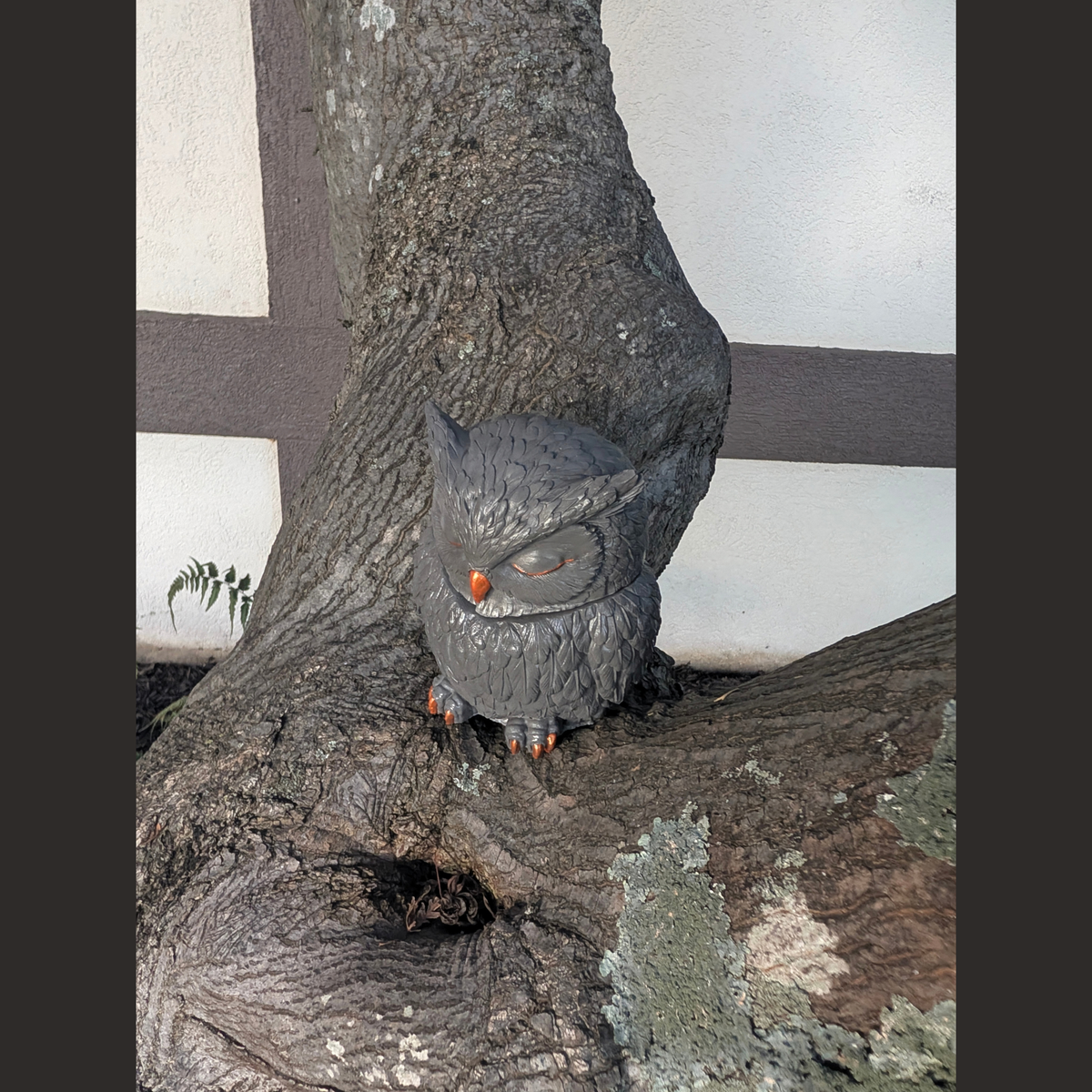 A small gray owl handcrafted vessel with orange feet and a yellow-orange beak perched on a large tree trunk outdoors against a white wall with dark trim.