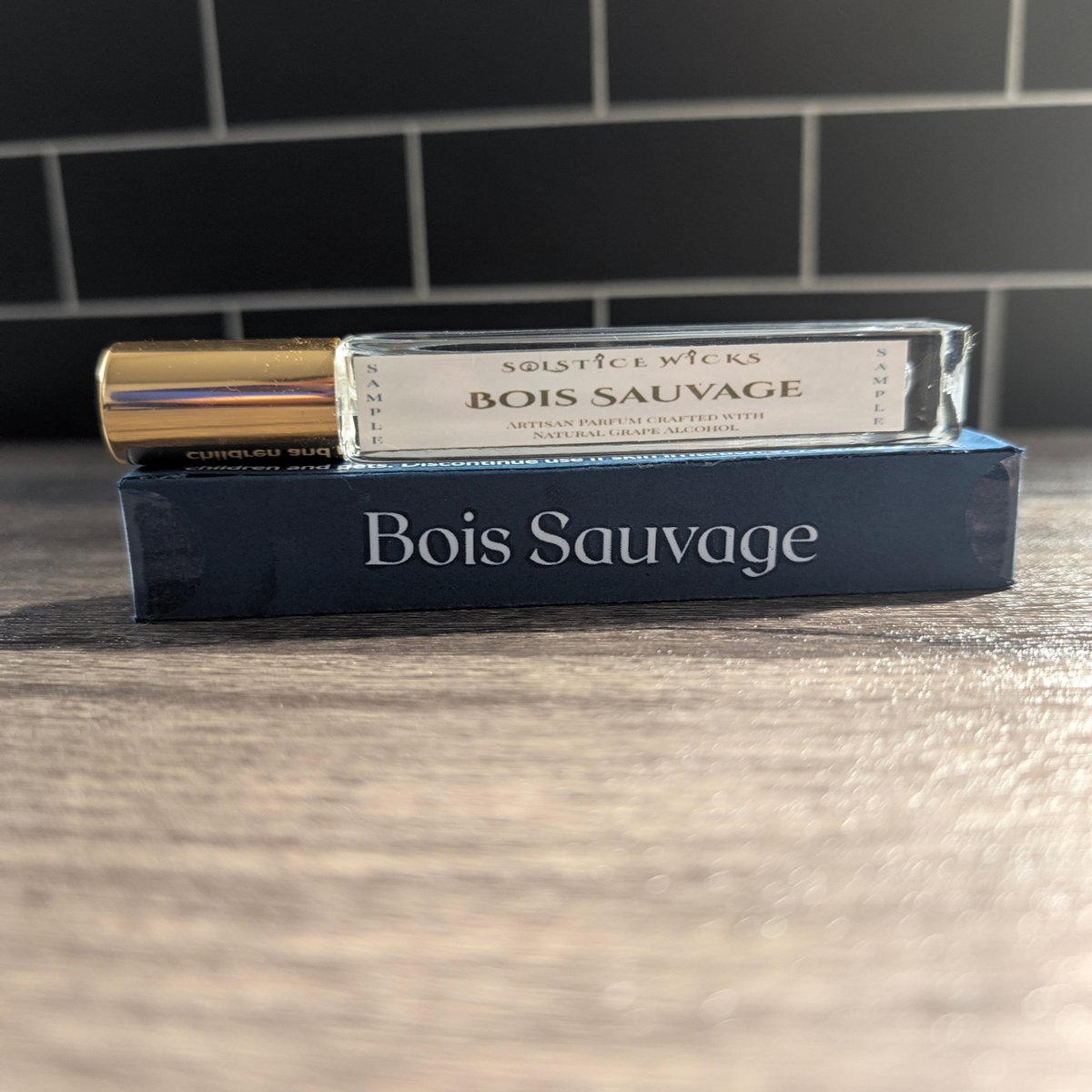 A glass To-Go spray bottle of Bois Sauvage by Solstice Wicks with a gold cap, resting on top of its dark blue cardboard packaging box, set on a wooden surface with a black tiled background.
