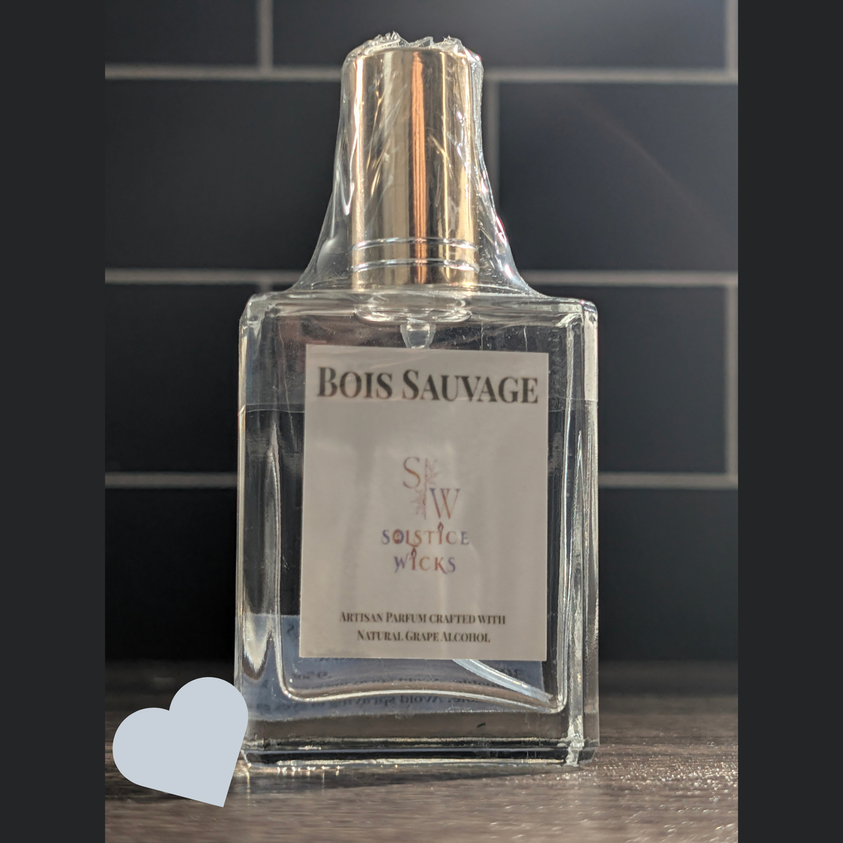 A bottle of Bois Sauvage perfume by Solstice Wicks, wrapped in protective plastic and featuring a white label, set against a dark tiled background.