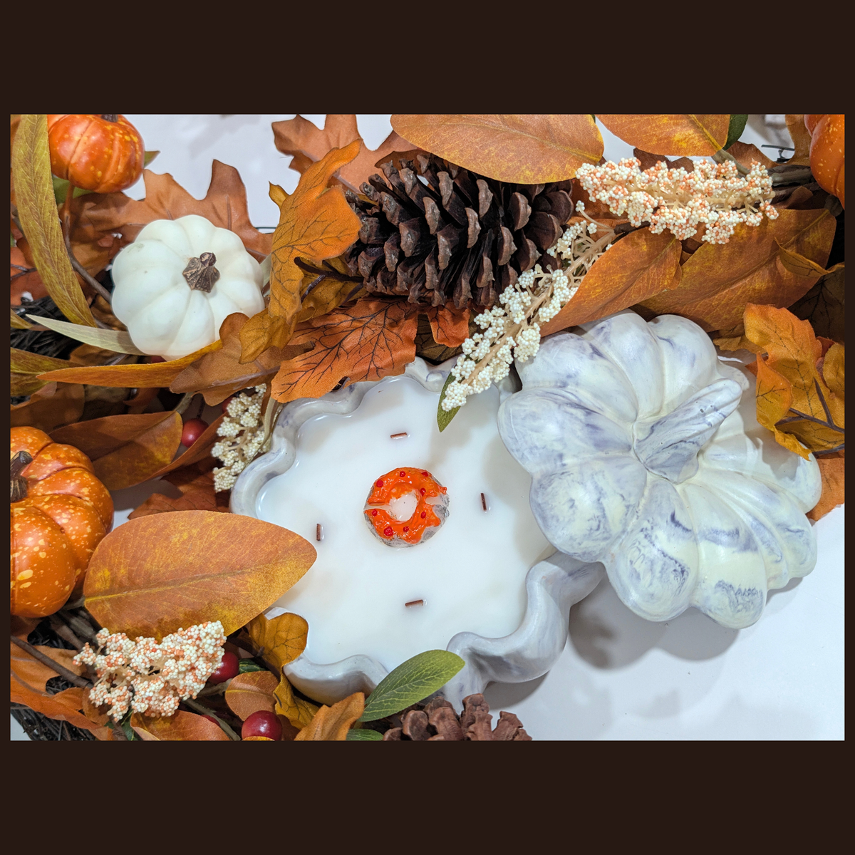 A handmade pumpkin candle arrangement featuring a white scalloped flower-shaped candle with a small cake  topper, surrounded by faux pumpkins, pinecones, and fall leaves. 