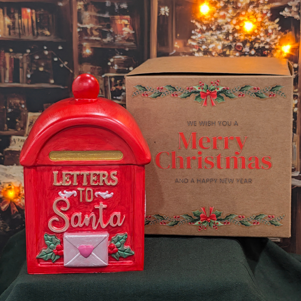 We have a red  mail vessel  with letters to Santa with a white envelope with a heart in front in the back have a brown box with Christmas decoration and letters saying we wish you a merry Christmas and a Happy New Year.  The box and vessel are in top of green mantel and Christmas photo as a background.