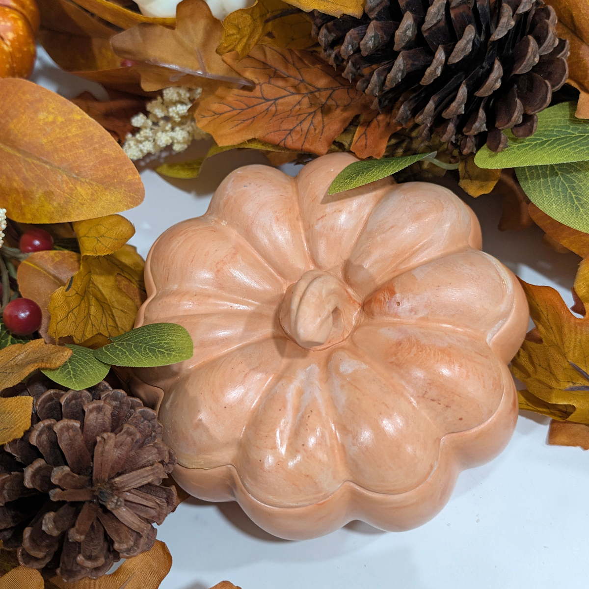 A Handmade pumpkin candle, surrounded by faux pumpkins, pinecones, and fall leaves. 