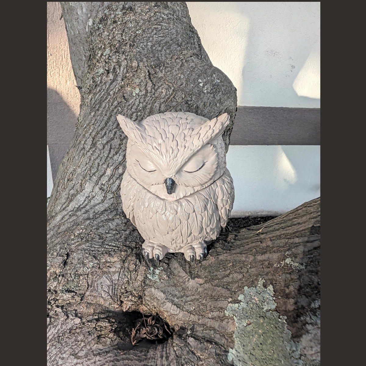 A small handcrafted owl figurine with closed eyes, perched on a textured tree trunk outdoors.
